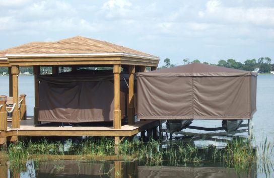 Two docked pontoon boats fully covered by Touchless Covers®; left boat under a roofed boat house, right boat elevated above water